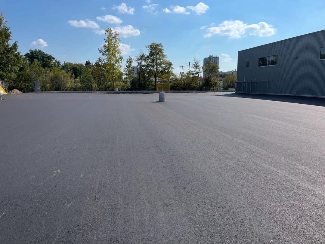 B9A58B48-62FC-408F-877C-02F64BA606E9 Freshly paved commercial parking lot beside a metal-clad building under a clear blue sky, with trees and utility infrastructure visible in the background.