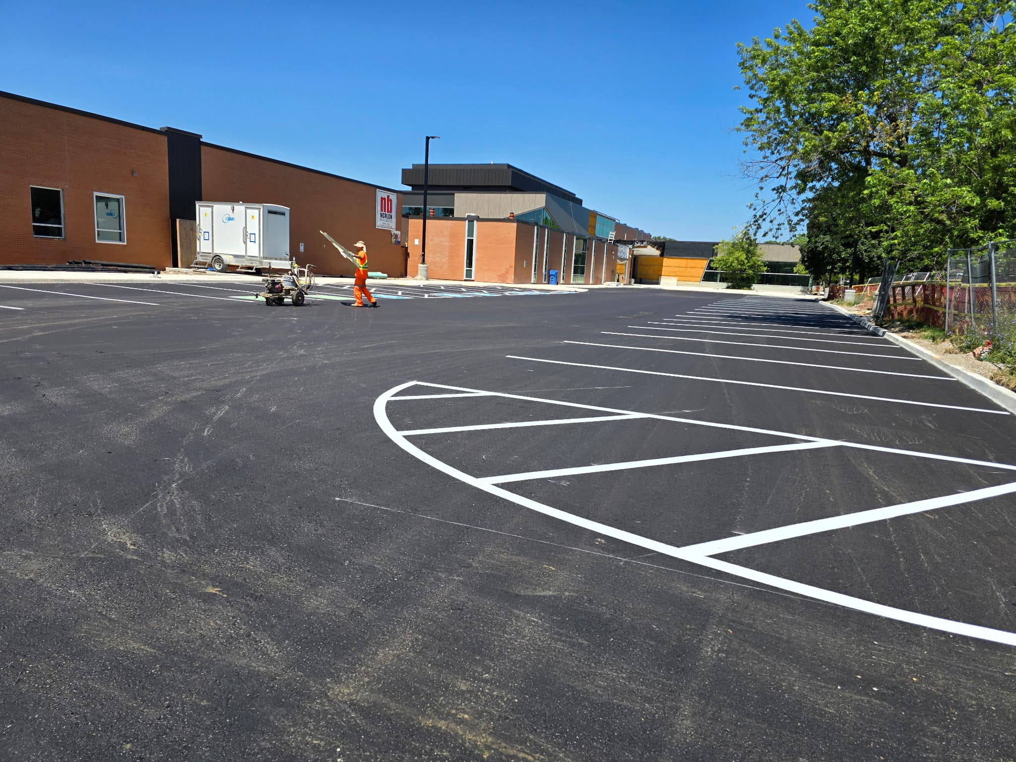 Freshly paved commercial parking lot in Kitchener, ON with newly painted white line markings. A crew member in safety gear operates a line striping machine under clear blue skies beside a red-brick retail building.