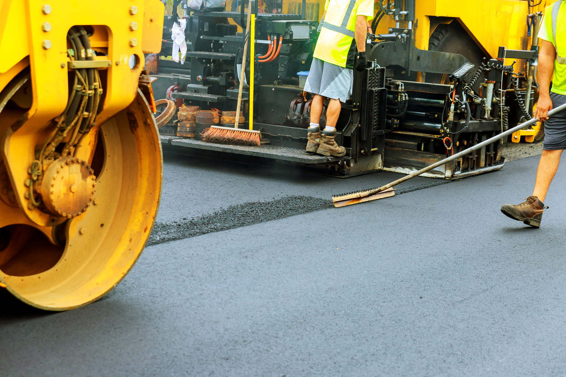 Workers performing asphalt paving using heavy machinery and hand tools, smoothing out a fresh layer of asphalt on a commercial roadway.
