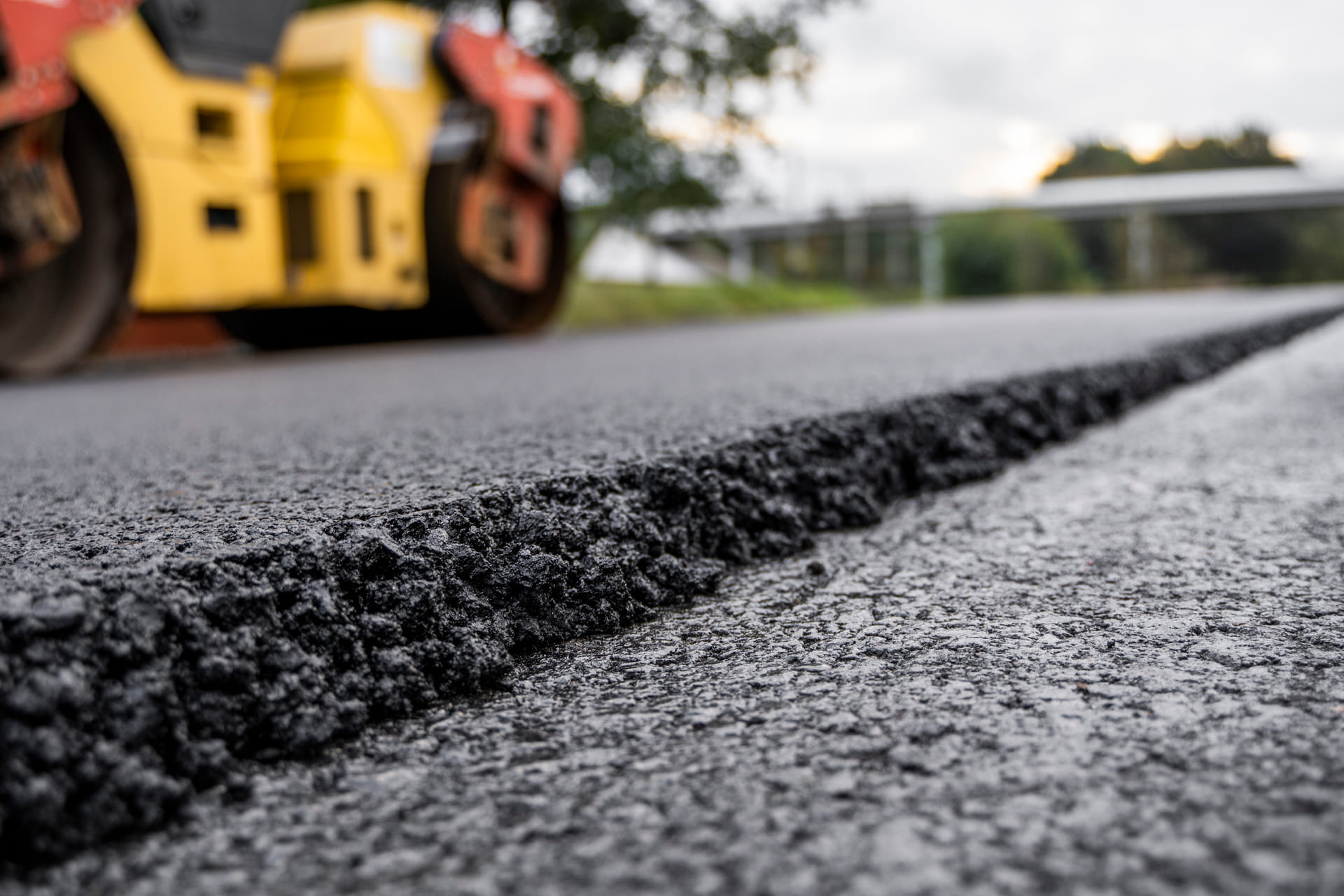 Close-up view of fresh asphalt during a roadway paving project, with a roller compactor in the background smoothing the new surface.