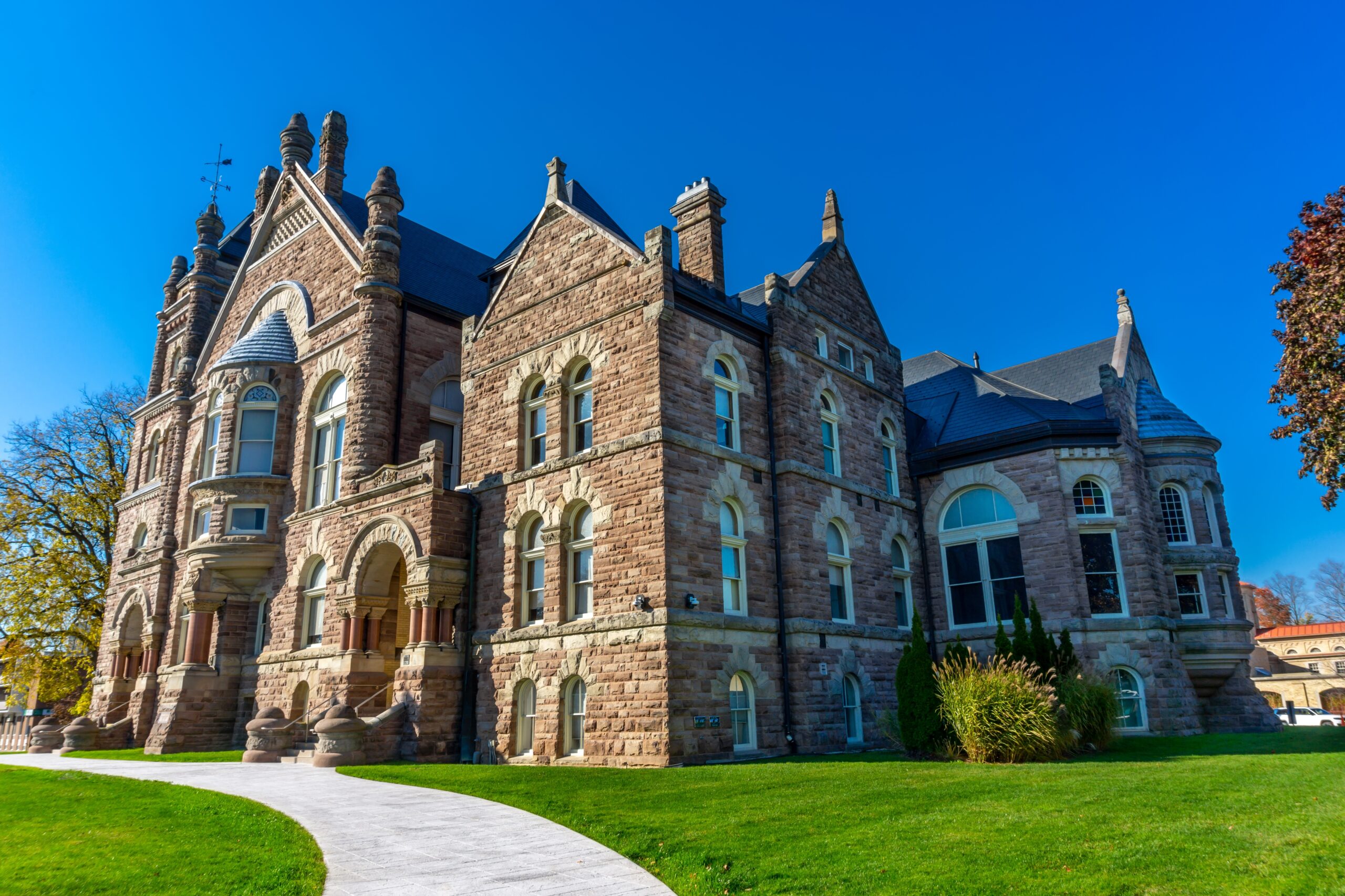 Historic sandstone building in Woodstock, Ontario under a clear blue sky, surrounded by green grass and a curved walkway leading to the entrance. This architectural landmark reflects the charm and heritage of the region.