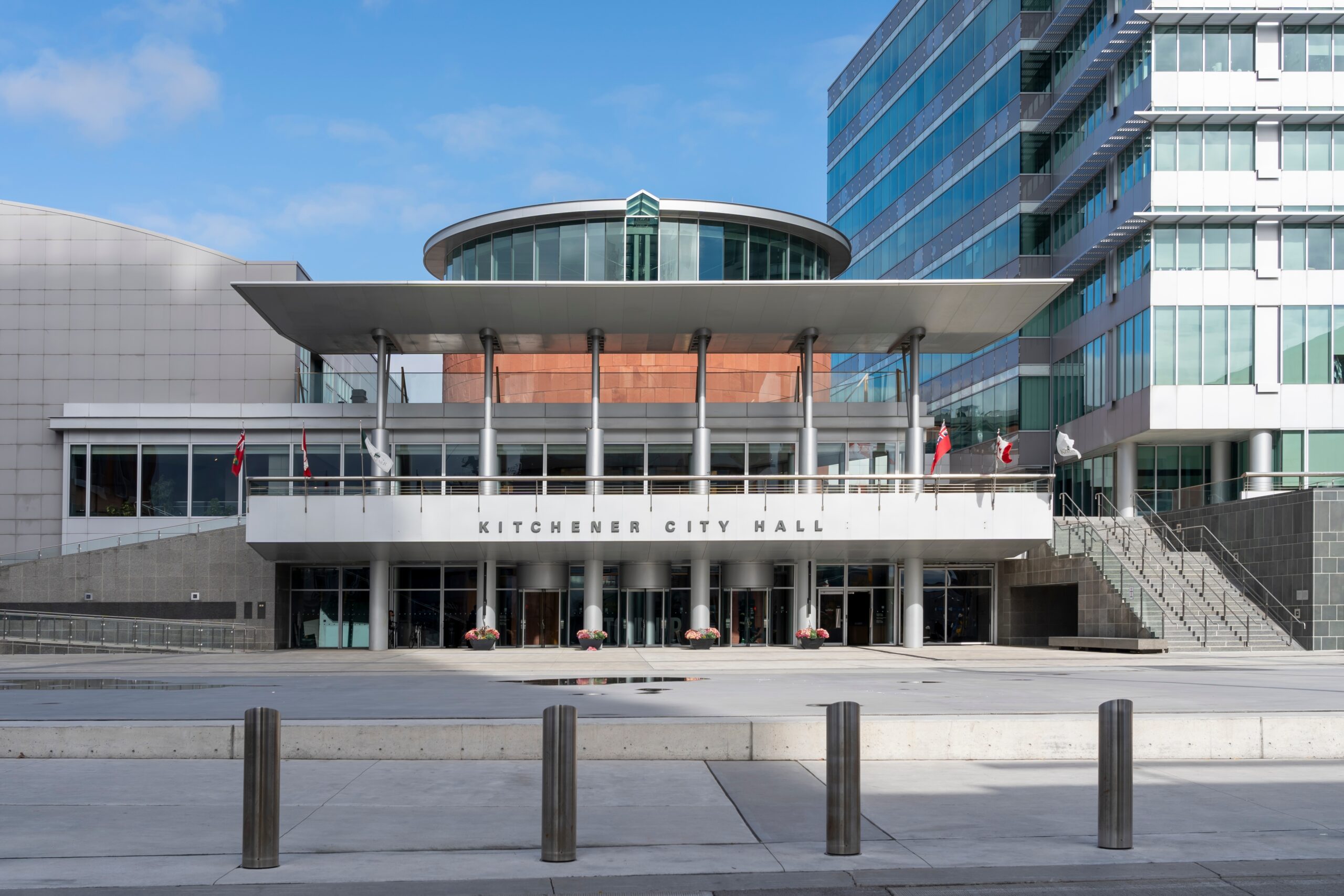 Modern exterior of Kitchener City Hall on a bright day, featuring glass walls, a circular copper structure, Canadian flags, and wide concrete steps in downtown Kitchener, Ontario.